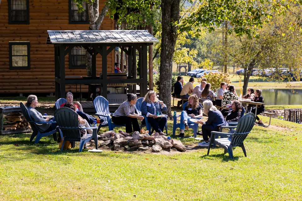 A circle of attendees in blue Adirondack chairs around a stone fire ring beside a log cabin