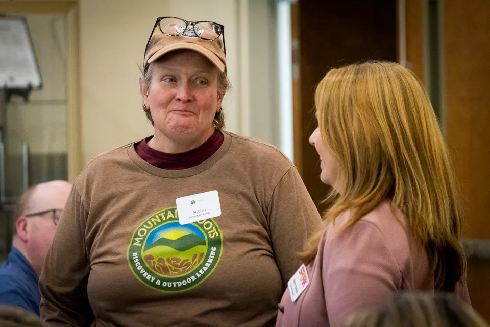 A woman in a ball cap and a brown T-shirt with a mountain logo smiles as she listens to another attendee.