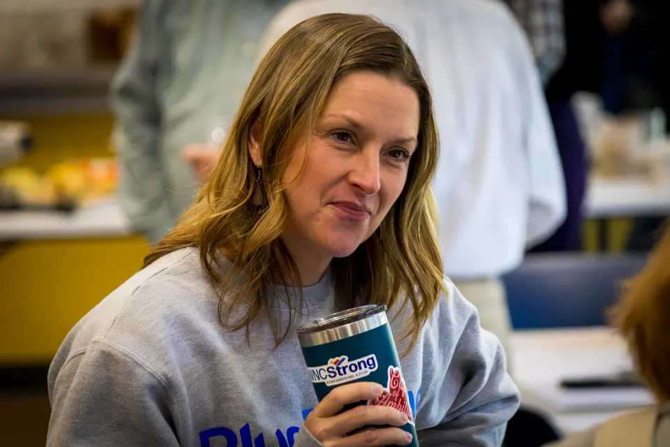 A woman in a gray sweatshirt holds an insulated tumbler and smiles at someone across the room.
