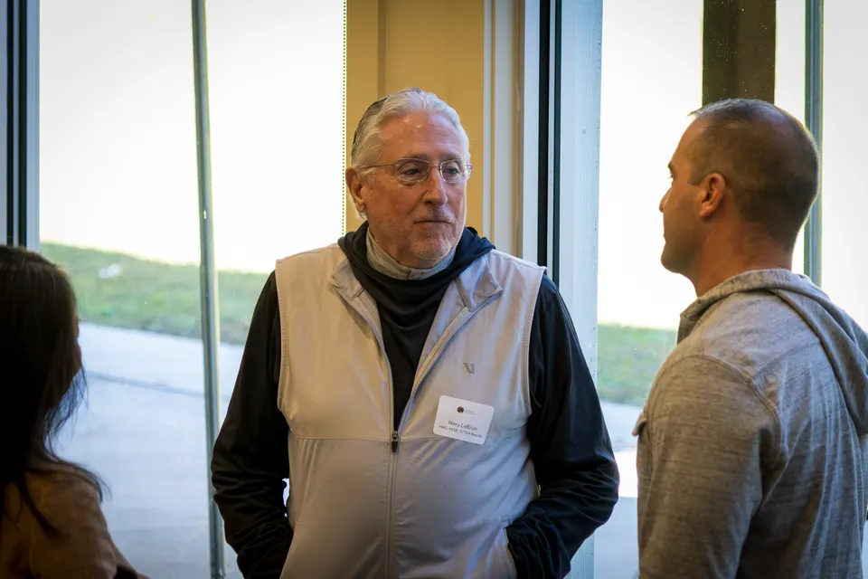 An older man in a vest talks with another attendee beside a tall window.