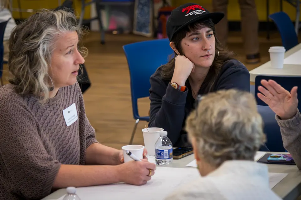 A woman listens closely at a table as the person beside her, in a black cap, makes a point.