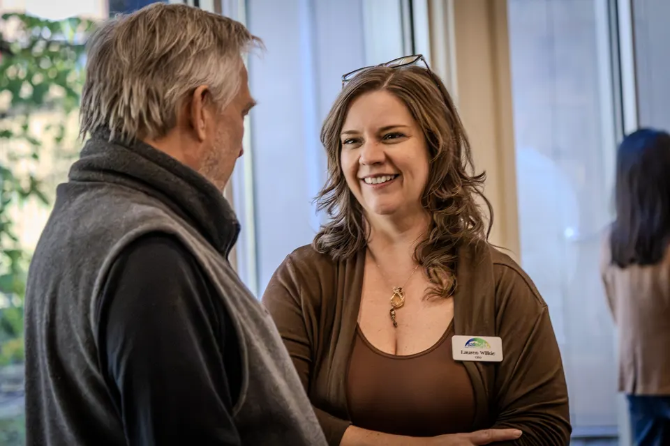 A man and a smiling woman in a brown sweater talk near the windows.