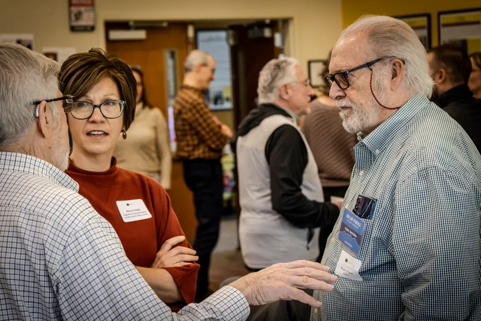 A crowded room of attendees mingling, a man with a white beard mid-conversation in the foreground.