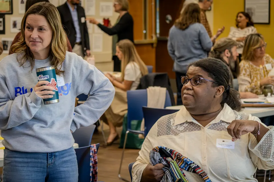 A woman in a gray sweatshirt stands among the tables, talking with an attendee seated beside a patterned bag.