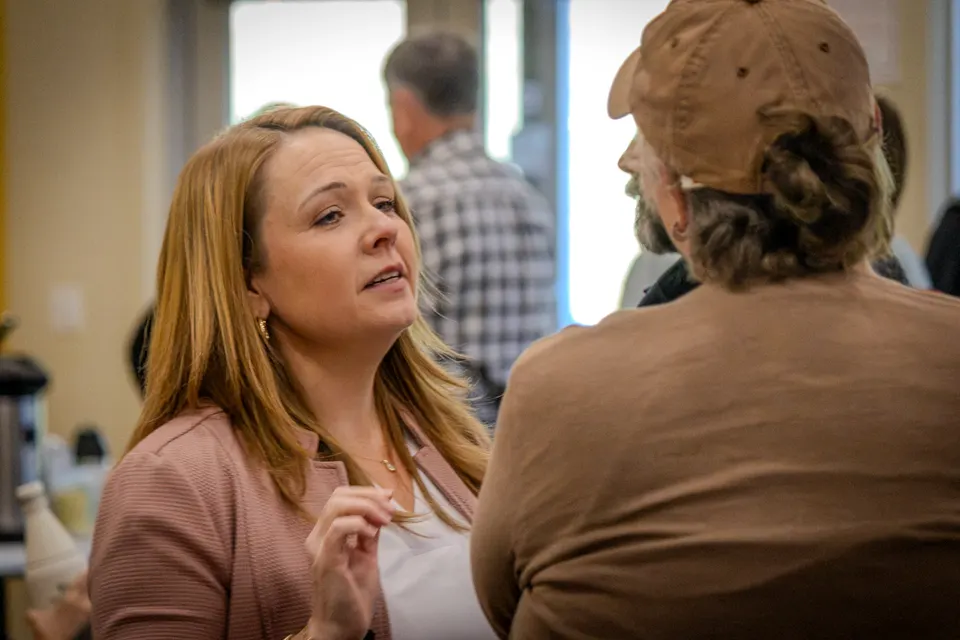 A woman in a pink cardigan speaks with an attendee in a tan cap.