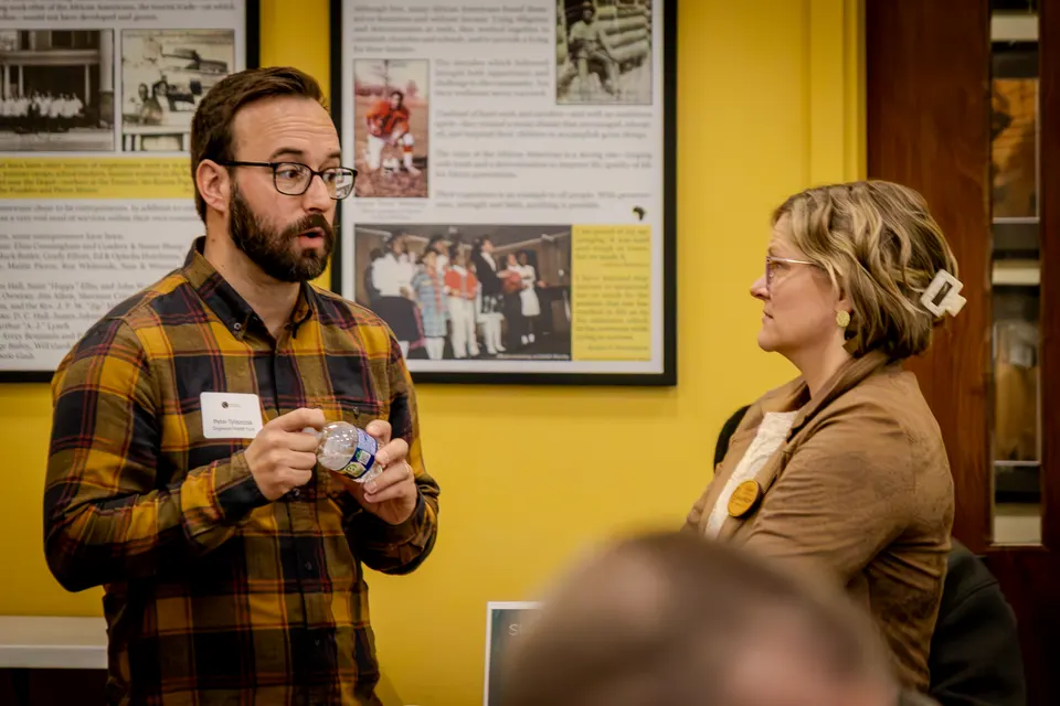A man in a plaid shirt holds a small item as he talks with a woman, framed photos on the yellow wall behind them.