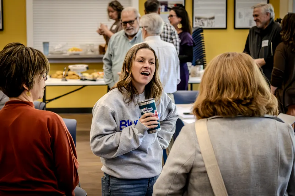 A woman in a gray sweatshirt laughs in the middle of the crowd near the refreshments table.